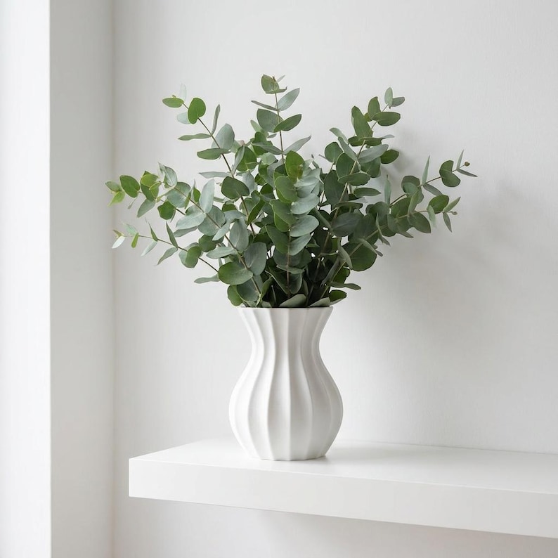 White matte textured fluted modern vase with green eucalyptus leaves on a white shelf against a white wall.