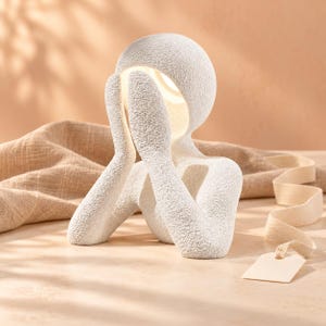 Matte textured off-white night light shaped like a seated figure with hands on its face, glowing soft warm white, on a peach-toned tabletop beside folded burlap fabric and a cream ribbon.