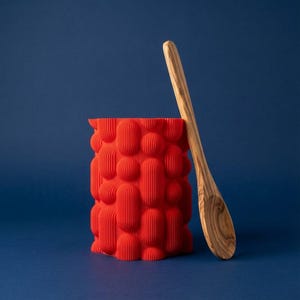 Textured matte red bubble vase with a wooden spoon leaning against it on a navy blue background.
