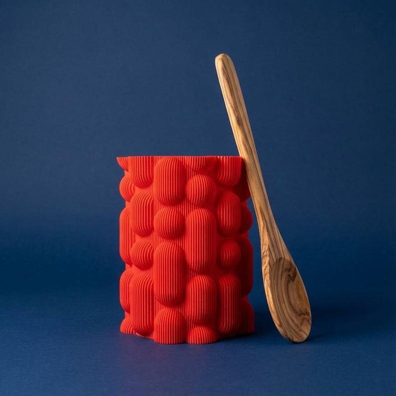 Textured matte red bubble vase with a wooden spoon leaning against it on a navy blue background.
