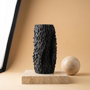 Puede incluir: Un jarrón negro texturizado con un diseño orgánico único. El jarrón se exhibe sobre una plataforma cuadrada de piedra beige, con un objeto esférico similar al mármol a la derecha. El fondo es de un tono cálido y neutro.