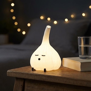 May include: A white, glowing, garlic-shaped lamp with a smooth, ribbed texture. The lamp features a simple, printed face with two horizontal lines for closed eyes and a small circle for a mouth. It has two small, dark, protruding feet at the base.
