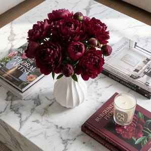 Burgundy peonies in a white matte textured fluted flower vase on a white-veined tabletop with stacked books, a candle and a burgundy book.