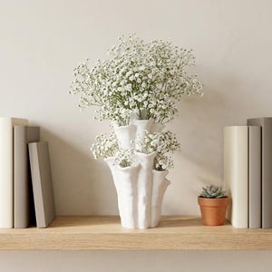 Matte white textured coral vase filled with white baby's breath on a shelf between two stacks of books, beside a small potted succulent. Dried flower vase about 7.2 in tall.