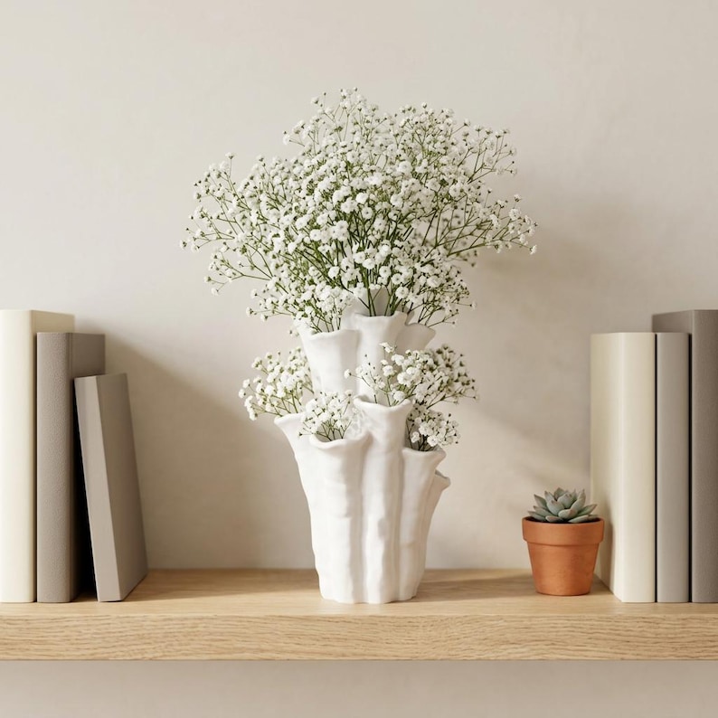 Matte white textured coral vase filled with white baby's breath on a shelf between two stacks of books, beside a small potted succulent. Dried flower vase about 7.2 in tall.