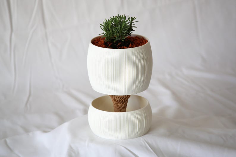 Textured matte white ribbed self watering pot holding a small green plant in reddish-brown soil, resting on a matching lower bowl against white fabric.