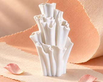May include: A white ceramic vase with a multi-tiered, organic structure resembling coral or ruffled flower petals. The object features several vertical, tubular openings of varying heights with wavy, scalloped edges and a smooth matte finish.
