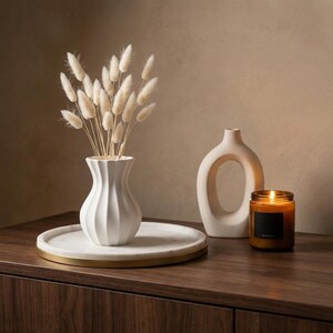 White matte textured fluted vase holding bleached dried grass plumes on a white tray atop a dark wood sideboard, with a lit candle and a loop-shaped vase nearby.