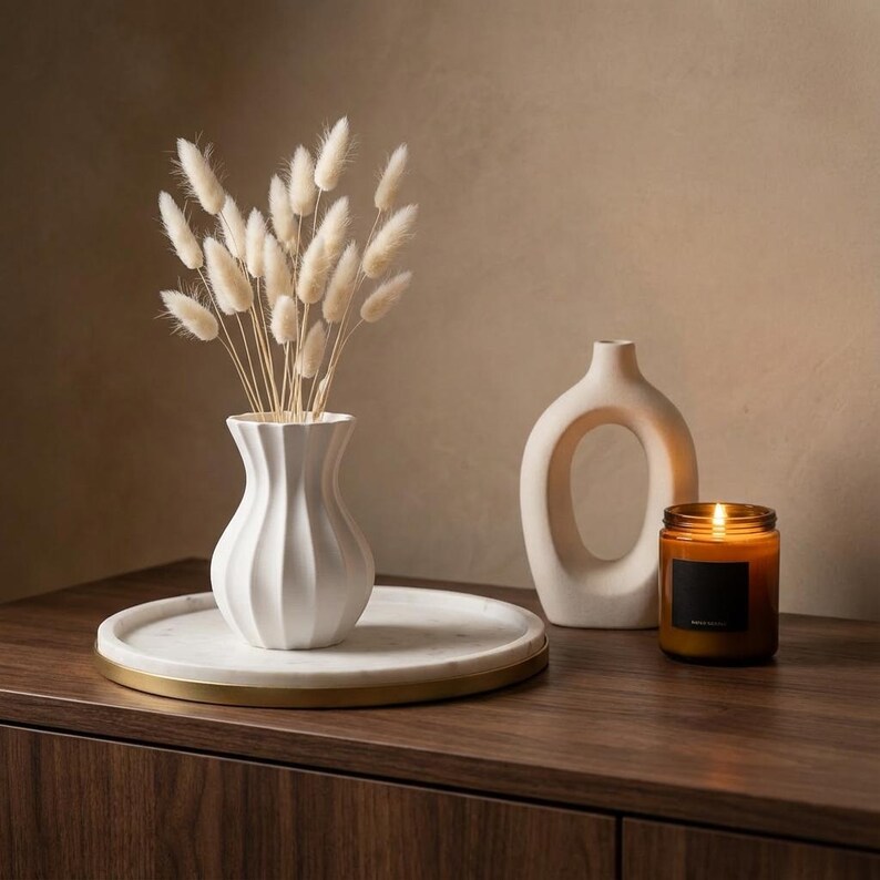 White matte textured fluted vase holding bleached dried grass plumes on a white tray atop a dark wood sideboard, with a lit candle and a loop-shaped vase nearby.