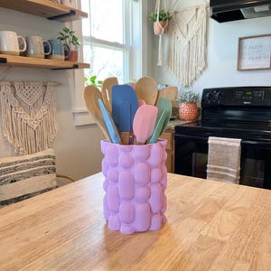 Textured matte lavender vase holding colorful spatulas and wooden spoons on a wooden kitchen table near a bright window.