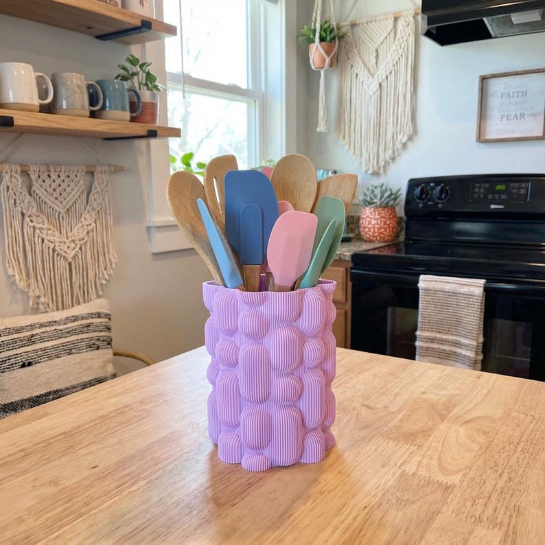 Textured matte lavender vase holding colorful spatulas and wooden spoons on a wooden kitchen table near a bright window.