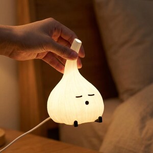 Hand holding a glowing garlic lamp by the stem, with a USB cable and bedding behind