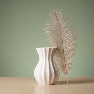 White matte textured fluted flower vase with a tall dried pampas plume leaning against it on a light surface, with a pale sage green background.