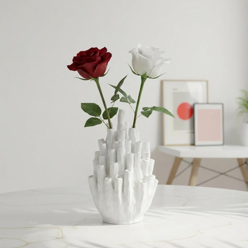 Matte white sculptural vase holding dark red flowers on a light gray surface with a minimal background.