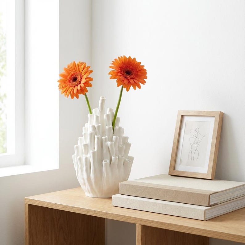 Matte white coral vase with orange flowers on a wooden table by a window, beside books, as a bud vase in japandi decor.