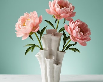 May include: A white ceramic vase with a textured, multi-tube structure resembling coral, holding three large, blooming pink peony flowers with green stems and leaves. The vase has a matte finish and multiple openings at varying heights.