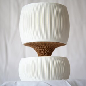 Textured matte white ribbed upper pot stacked over a lower bowl, joined by a brown cone-shaped core, against a white fabric backdrop; a minimal self watering pot.