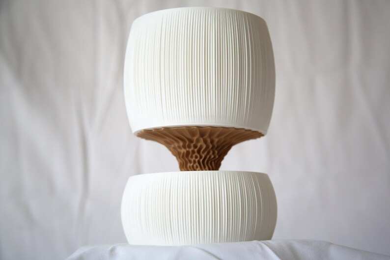 Textured matte white ribbed upper pot stacked over a lower bowl, joined by a brown cone-shaped core, against a white fabric backdrop; a minimal self watering pot.