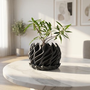 Op de afbeelding: Een donkergrijze, getextureerde, decoratieve plantenbak met een uniek golfachtig ontwerp, met een kleine groene plant. De plantenbak staat op een tafel met een marmeren blad. De plant heeft lange, slanke groene bladeren.
