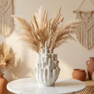 Matte white textured bud vase holding dried pampas grass on a light surface, with a boho wall hanging in the background, in warm japandi decor styling.