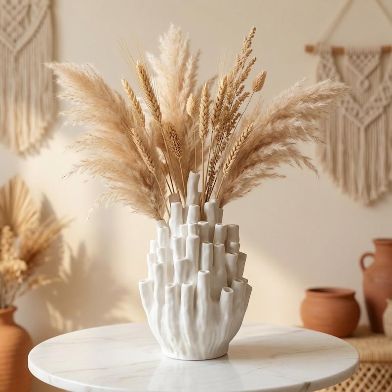 Matte white textured bud vase holding dried pampas grass on a light surface, with a boho wall hanging in the background, in warm japandi decor styling.