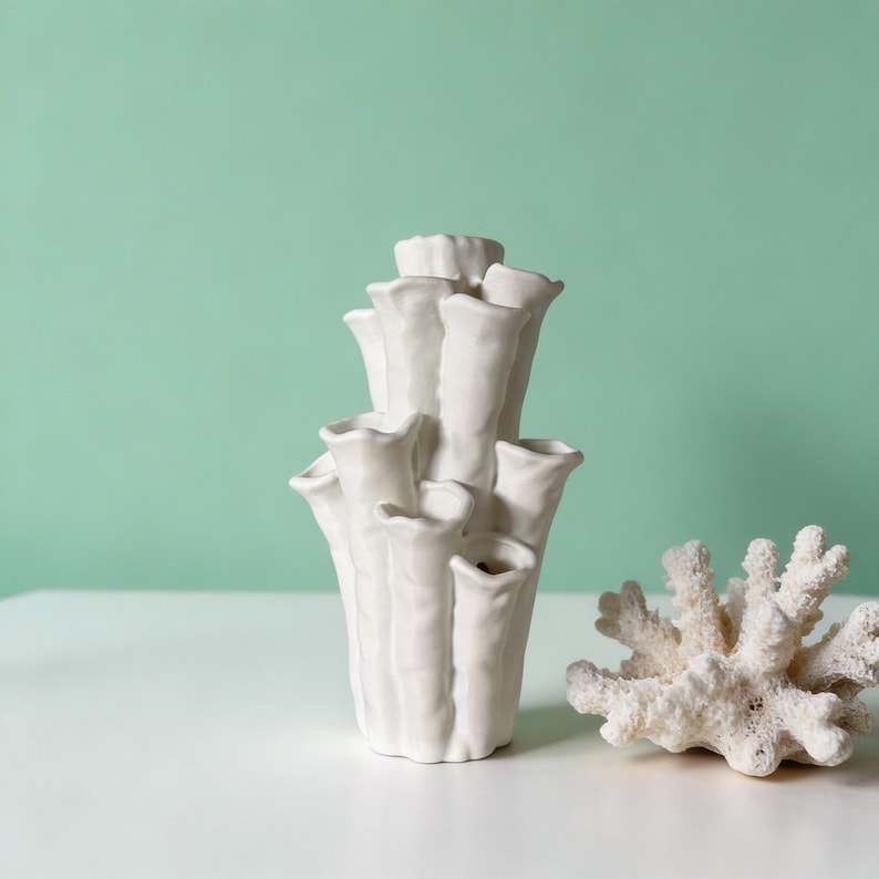 Matte white textured coral vase with several branching tubular openings, next to a piece of white dried coral on a smooth surface. Sculptural vase about 7.2 in tall and 3.9 in wide.
