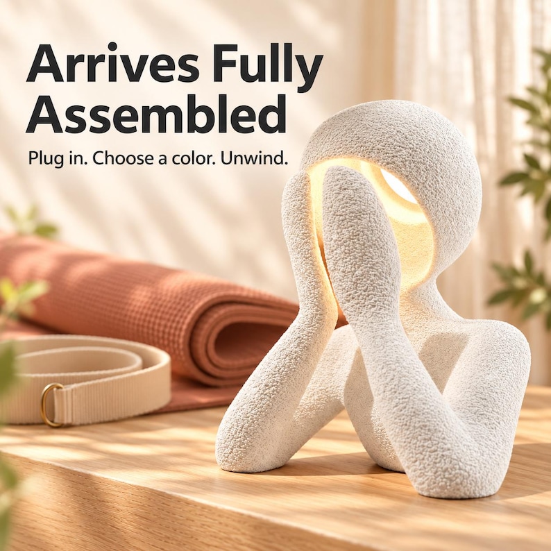 Matte textured white night light figure glowing warm yellow-white on a wooden shelf beside a rust-colored rolled mat, with text reading Arrives Fully Assembled.