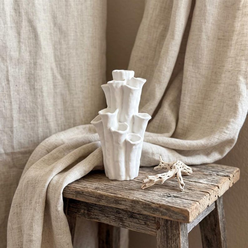 Matte white textured coral vase standing on a small wood table in front of a draped beige curtain. Sculptural vase for home decor, about 7.2 in tall and 3.9 in wide.
