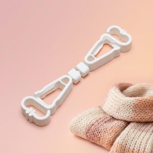 May include: A white plastic scarf hanger with a unique design, featuring two angled clips connected by a series of small blocks. A folded, knitted scarf in shades of pink, cream, and peach is next to the hanger, suggesting its use for storing scarves.
