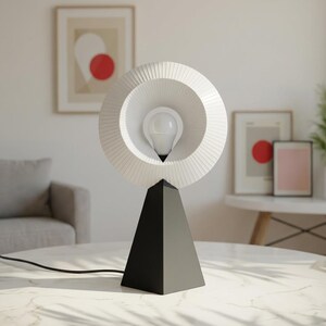 Modern table lamp with a pleated matte white ring shade around a bulb, on a matte black pyramid base on a white-veined tabletop, with framed art and a round side table behind.