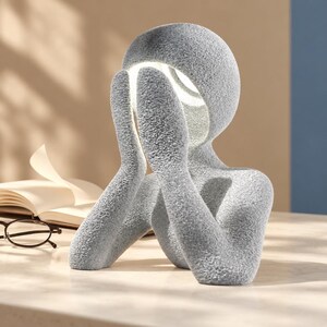 Gray matte textured abstract lamp shaped like a figure with hands over its face, glowing white from the head, on a light tabletop beside an open book with a blue wall behind.