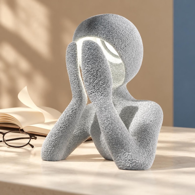 Gray matte textured abstract lamp shaped like a figure with hands over its face, glowing white from the head, on a light tabletop beside an open book with a blue wall behind.