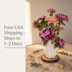 Matte white textured coral vase holding pink and mauve dianthus flowers on a round wood slice, with a "Free USA Shipping" text banner at left. Dried flower vase style, about 7.2 in tall.