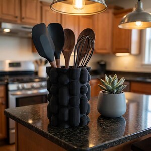 Textured matte black vase holding black and wooden utensils on a dark kitchen counter next to a small potted succulent.