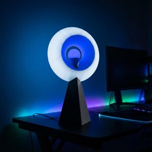 Small table lamp with a pleated white ring shade glowing bright blue, on a matte black base on a dark desk with a monitor and green and purple accent lights.