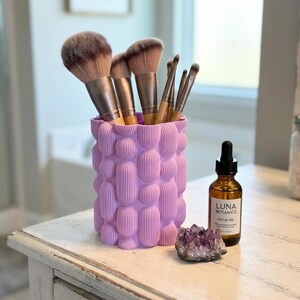 Textured matte lavender vase holding makeup brushes on a worn white vanity beside a dropper bottle labeled "LUNA" and a purple crystal.