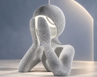 Puede incluir: Una escultura gris texturizada de una figura con cabeza redondeada y brazos elevados hacia el rostro. La figura cuenta con una fuente de luz integrada en la cabeza, que proyecta un suave resplandor tras las manos e ilumina la cavidad interior.
