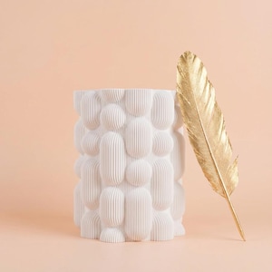 Matte white vase with a chunky bubble texture beside a gold feather against a peach background, a unique vase for modern shelves.