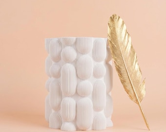 May include: A white, textured vase with a bubble-like design. A gold-colored feather leans against the vase. The vase has a cylindrical shape and is set against a peach-colored background.
