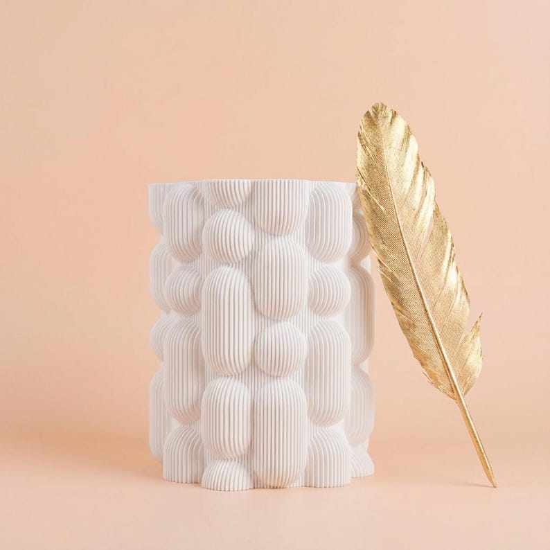 Matte white vase with a chunky bubble texture beside a gold feather against a peach background, a unique vase for modern shelves.