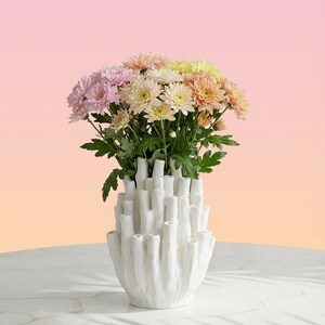 Matte white sculptural vase holding pink and yellow flowers against a pink and peach gradient background.