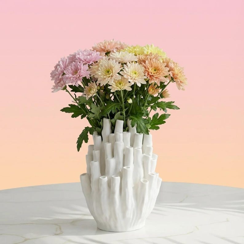 Matte white sculptural vase holding pink and yellow flowers against a pink and peach gradient background.