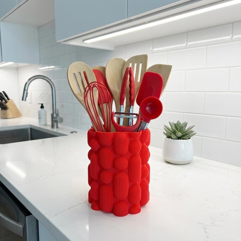 Textured matte red vase holding kitchen utensils on a white-veined tabletop counter beside a small potted succulent.