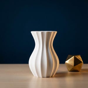 White matte textured fluted modern vase with a wavy silhouette on a wooden surface next to a gold geometric object, against a deep navy blue background.