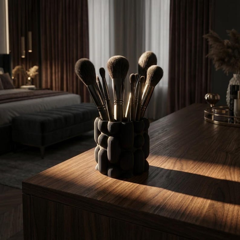 Textured matte black vase holding makeup brushes on a dark wooden table in a moody bedroom with warm sunlight.