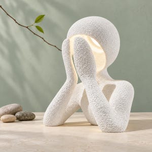 May include: A white table lamp shaped like a stylized human figure with a textured, granular surface. The figure has its hands raised to its face, and the head is a rounded dome that emits a soft, warm light from the interior cavity.