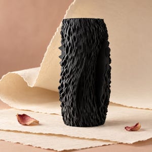 May include: A cylindrical black vase featuring a highly textured, vertical wavy surface pattern. The exterior is composed of numerous thin, repeating ridges that create a deep, rippled effect across the entire height of the object.