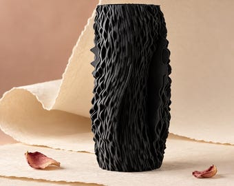 May include: A cylindrical black vase featuring a highly textured, vertical wavy surface pattern. The exterior is composed of numerous thin, repeating ridges that create a deep, rippled effect across the entire height of the object.