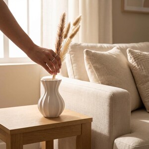 A hand arranges dried pampas grass in a white matte textured fluted minimalist vase on a wooden side table beside a beige sofa with cushions.