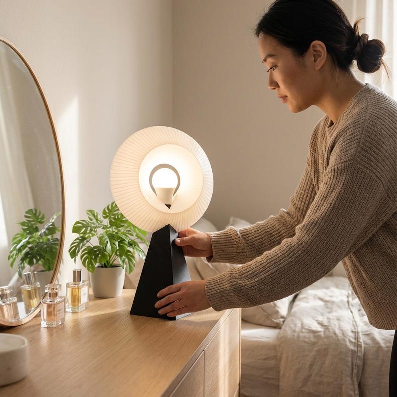 Woman adjusting a small table lamp with a pleated matte white ring shade glowing warm white and a matte black pyramid base on a wooden dresser in a bedroom.